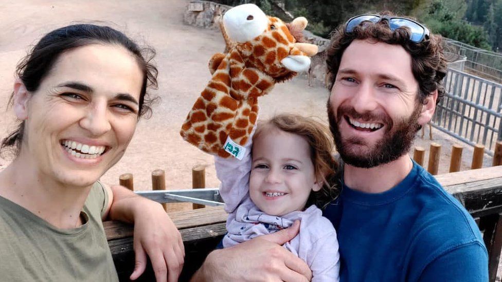 A photo of Jordan Roman-Gat, her husband Alon and three-year-old daughter Gefen, smiling as the child holds up a giraffe glove puppet