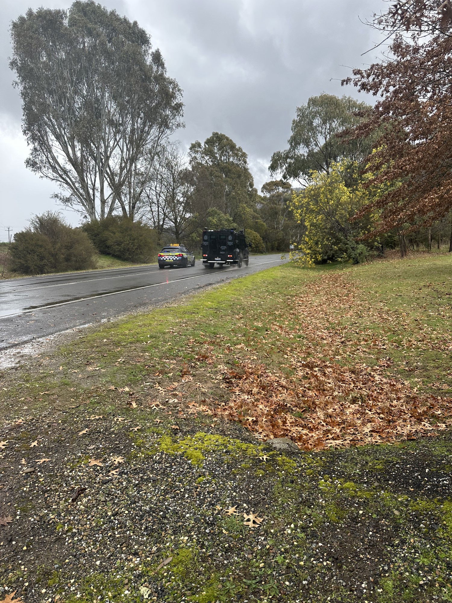 A dark fortified vehicle passes a police car on a wet rural road.
