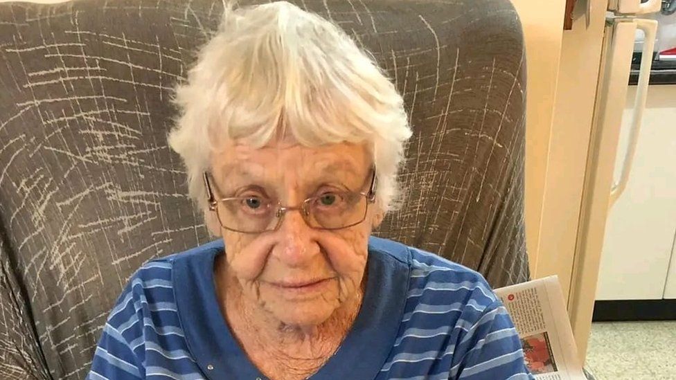 Family photo of Ditza Heiman, a white-haired woman with glasses, sitting in a chair looking up at the camera
