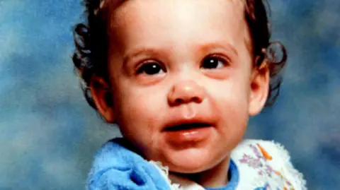 Family photo Picture of Katrice Lee. She has brown curly hair, brown eyes and is wearing a blue and white top in front of a blue background