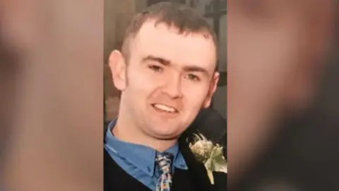 Family handout A man with short brown hair, wearing a black suit and blue shirt. The sides of the image are blurred. 