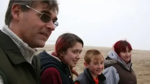 BBC Paul Jones on the left has dark hair and wears dark glasses, a a light coloured roll-neck jumper under a dark coat. To his right is a young girl with long brown hair and a dark hooded body-warmer and red jumper. To her right is a young boy with fair hair and dark body-warmer and red top. To his right is a woman with long dark hair, a green top under a dark body-warmer.
