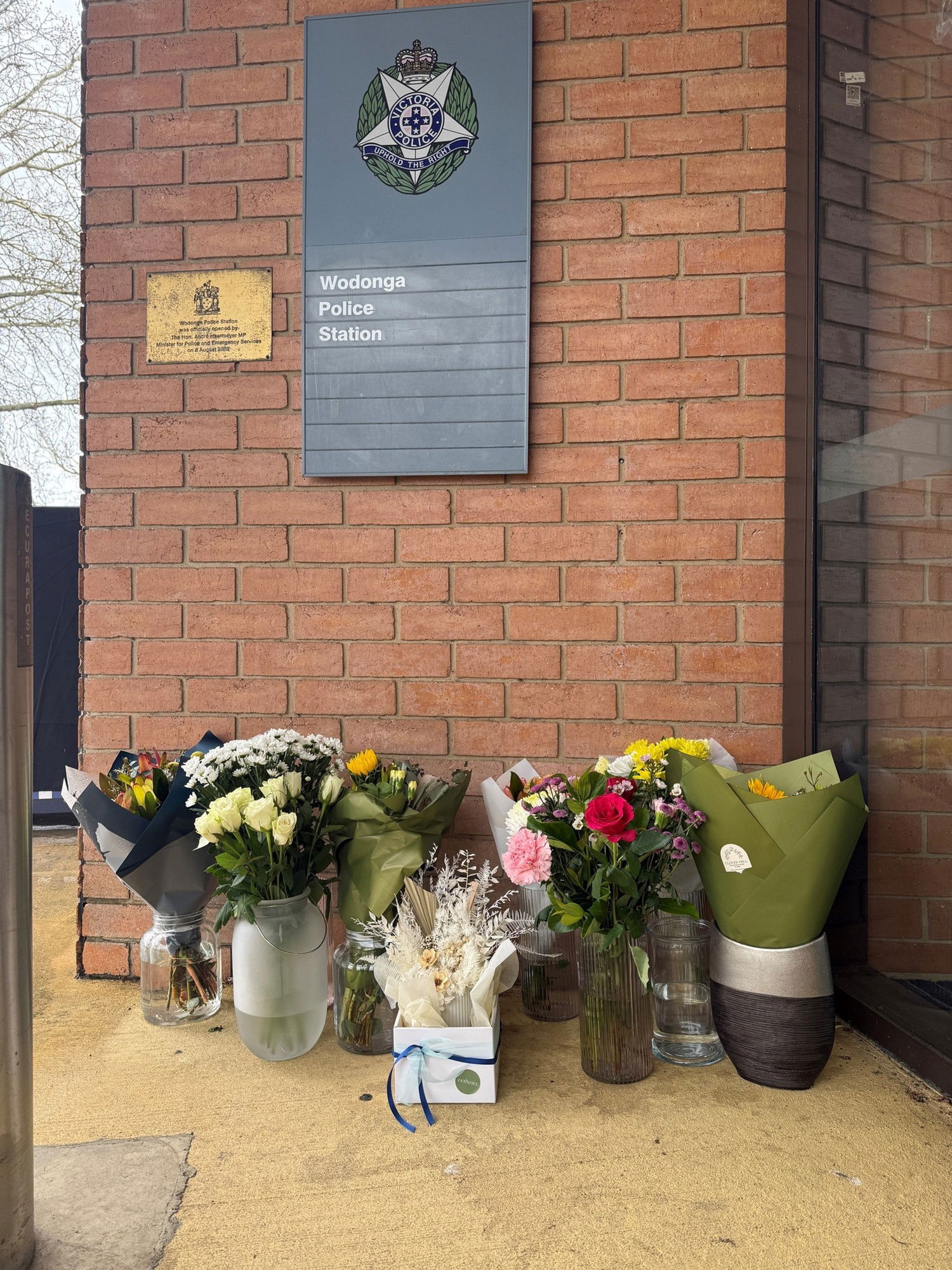 Bouquets of flowers in boxes and vases stand against a brick wall with a sign saying Wodonga Police Station.
