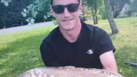 Family Louis Price is wearing a black T-shirt and dark sunglasses. He is holding a large fish close to the camera and smiling.